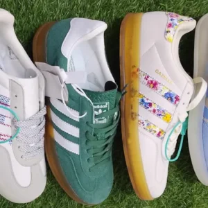 Cover image showing four pairs of Adidas Gazelle sneakers in Nairobi Kenya. Variants include white with multicolored stitched stripes, dark green with white stripes, white with floral patterned stripes, and light gray with pastel blue stripes. All feature gold 'GAZELLE' branding, gum soles, and classic retro design.